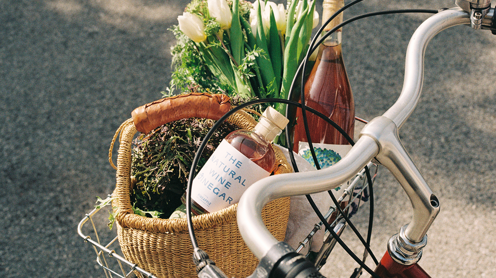 A bottle of The Natural Wine Vinegar by Pineapple Collaborative and Martha Stoumen in a bike basket, with a bottle of wine and a bouquet of tulips